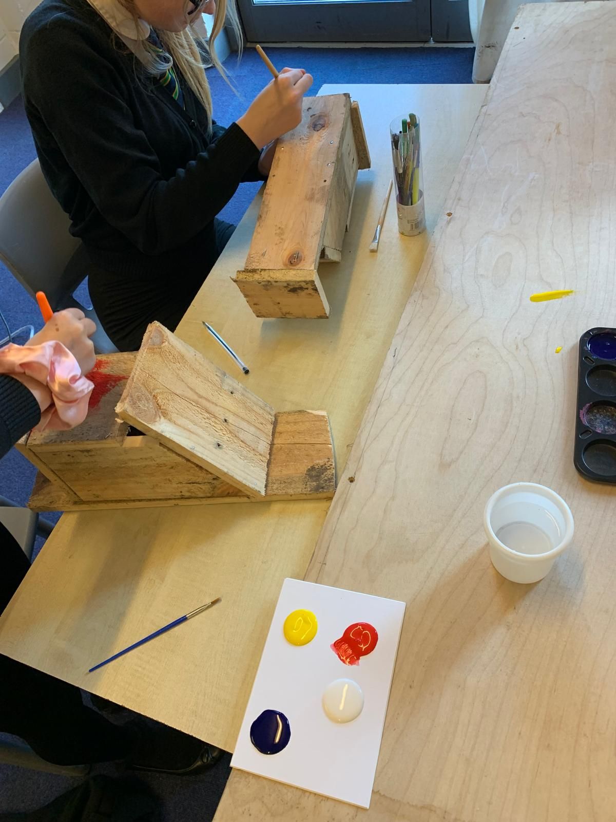 A person is sitting at a table painting a wooden birdhouse.