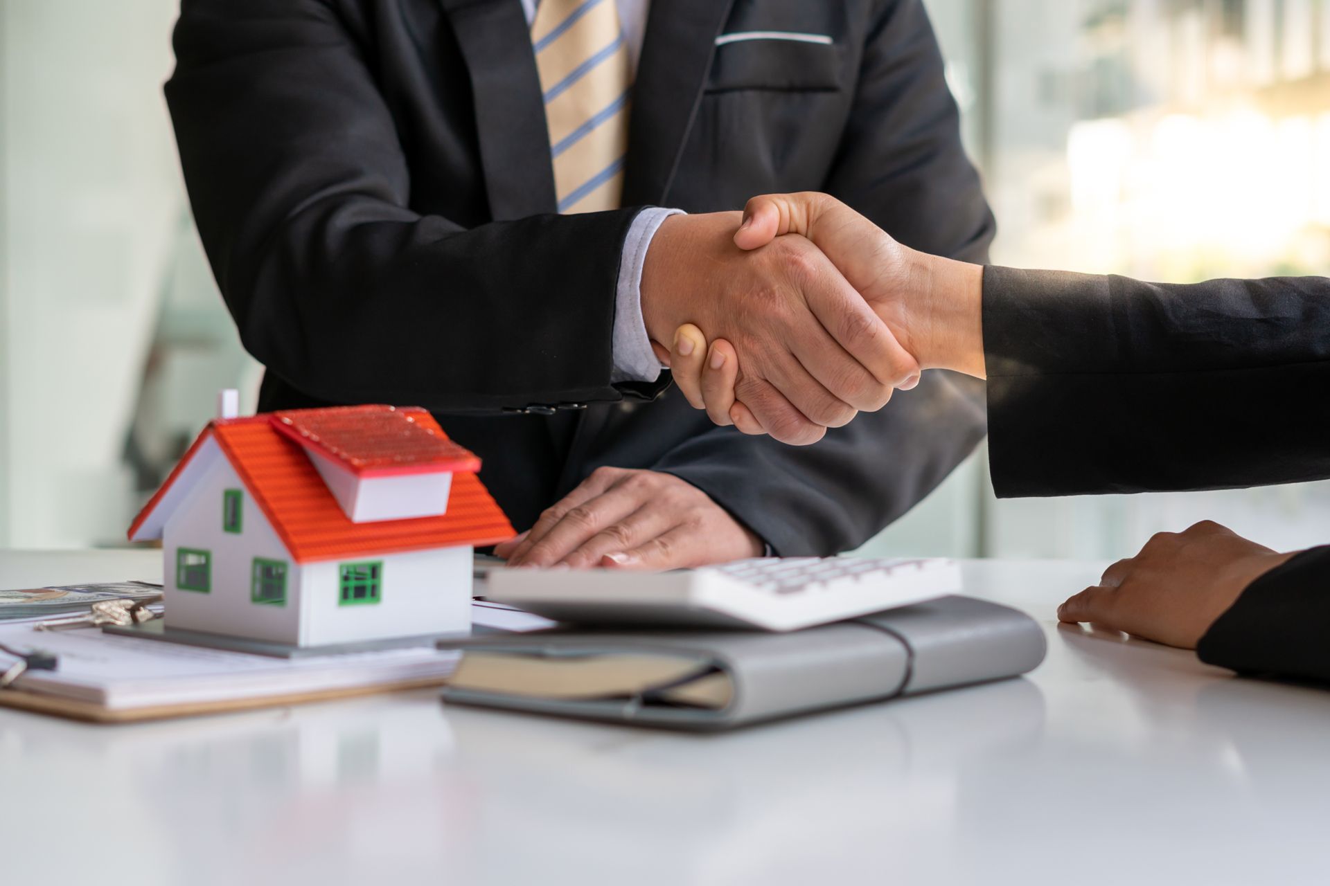 Two people in business attire shake hands over a desk with a small model house, a calculator, and documents.