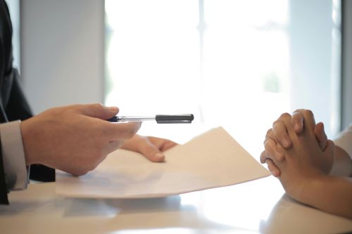Person in suit holding pen over paperwork, across from person with hands clasped. White interior, natural light