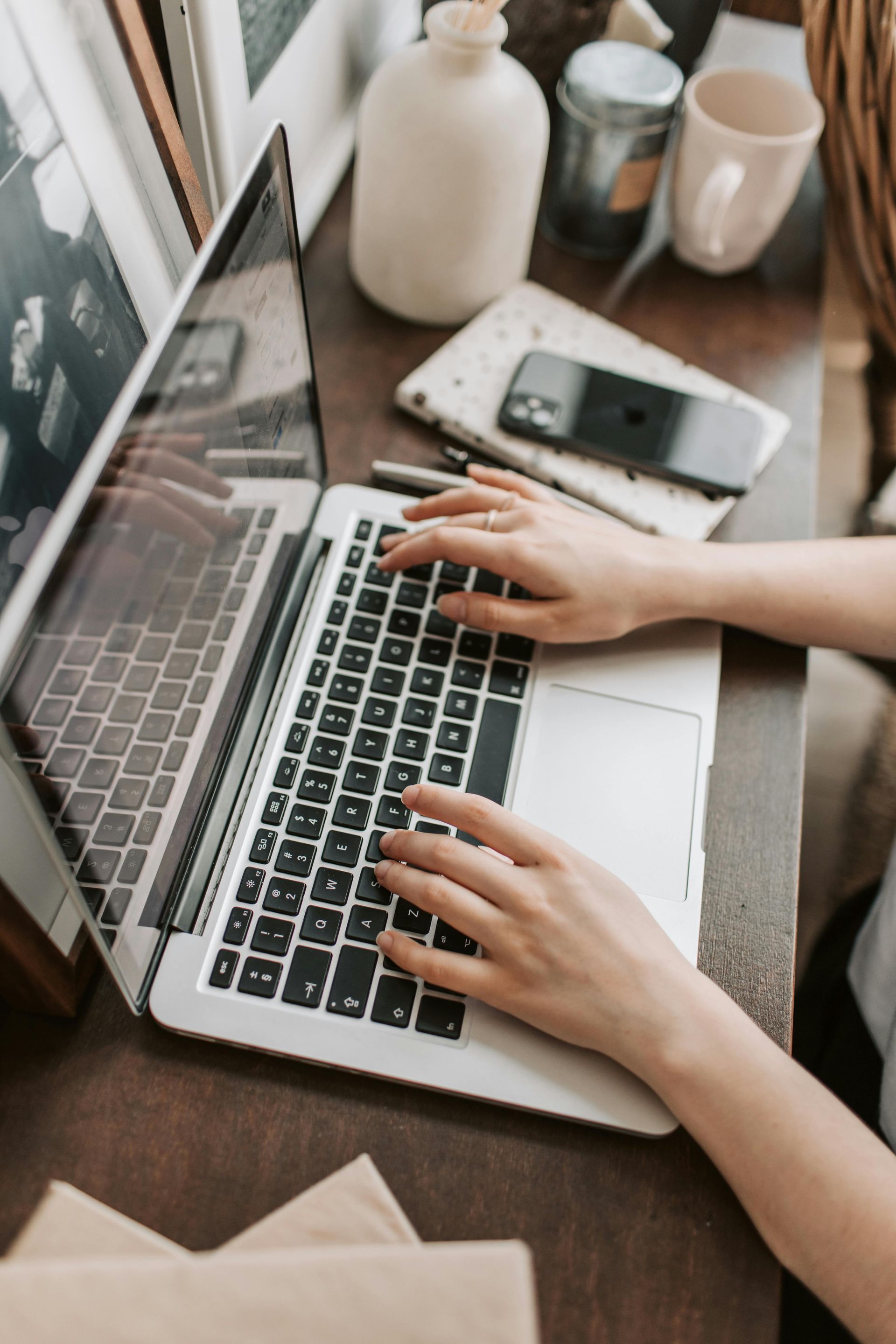 Woman typing on a laptop at a desk, with a phone, notepad, and coffee cup nearby