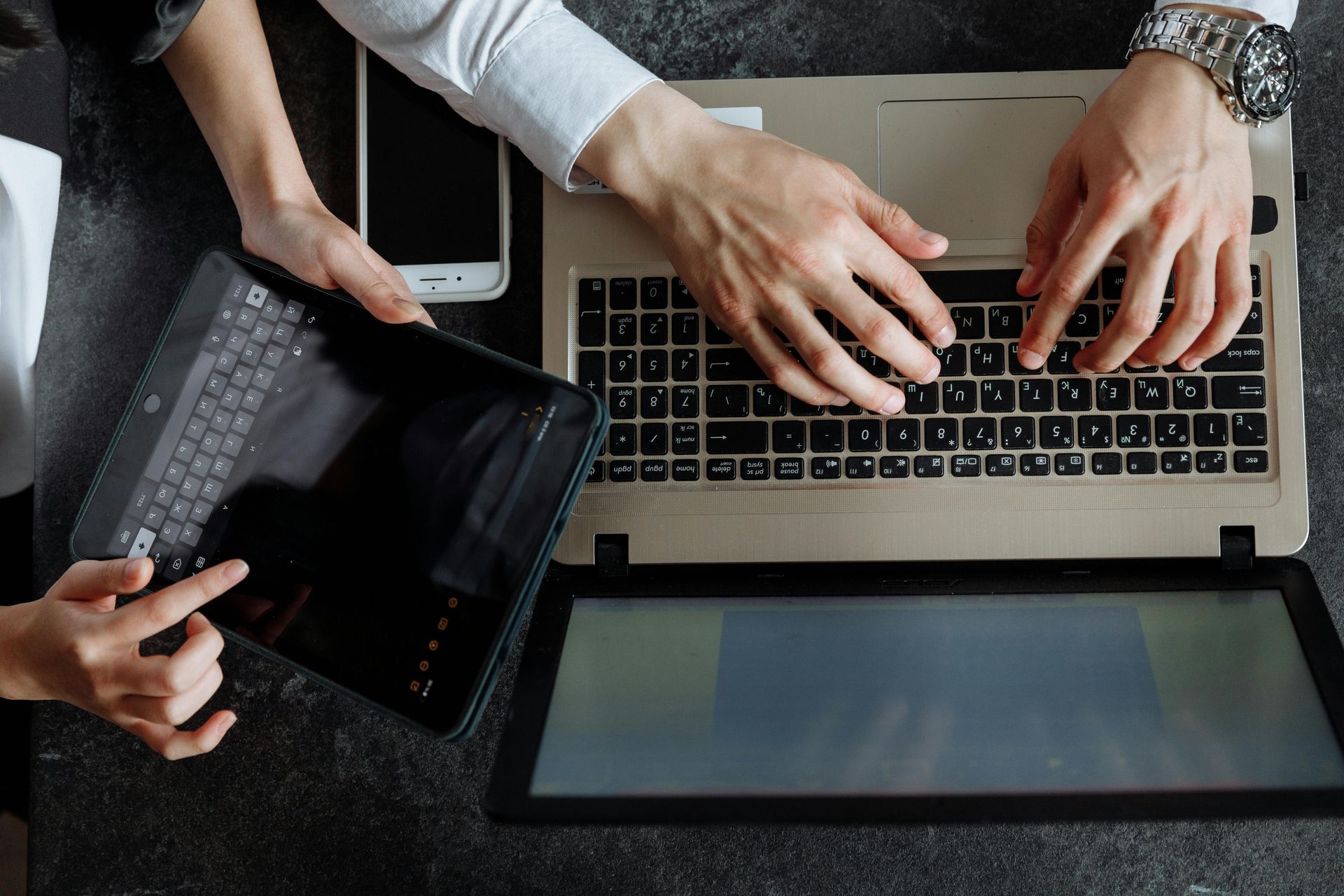 Two people work at a desk, one typing on a laptop and the other operating a tablet next to a smartphone.