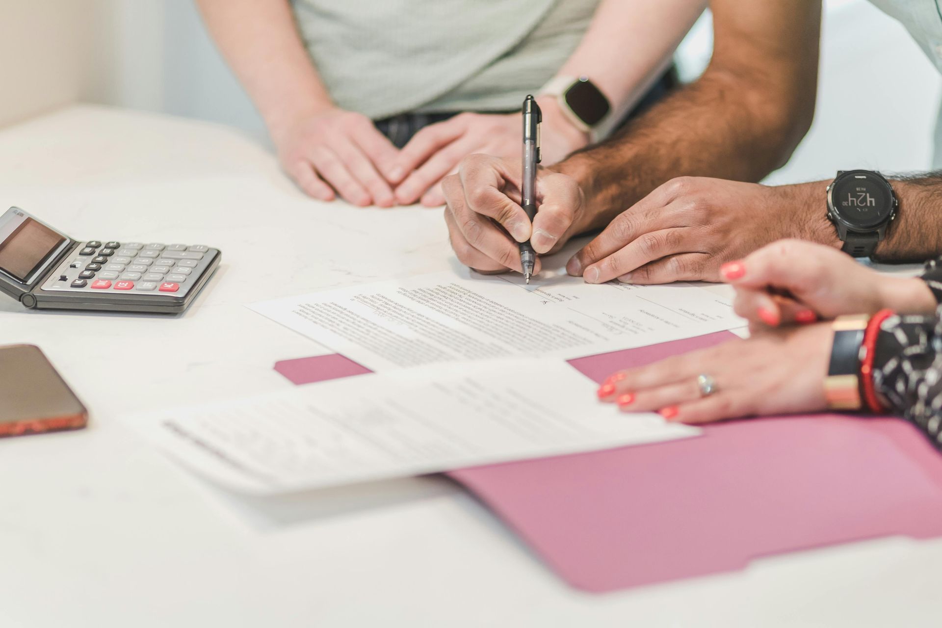 Hands signing a document on a white table; a calculator, phone, and pink folder are also present