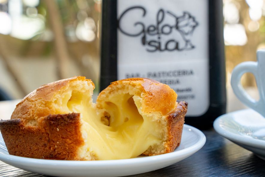 Un pasticcino alla crema pasticcera morso su un piatto bianco accanto a una bottiglia di vino e una tazza di caffè su un tavolo.