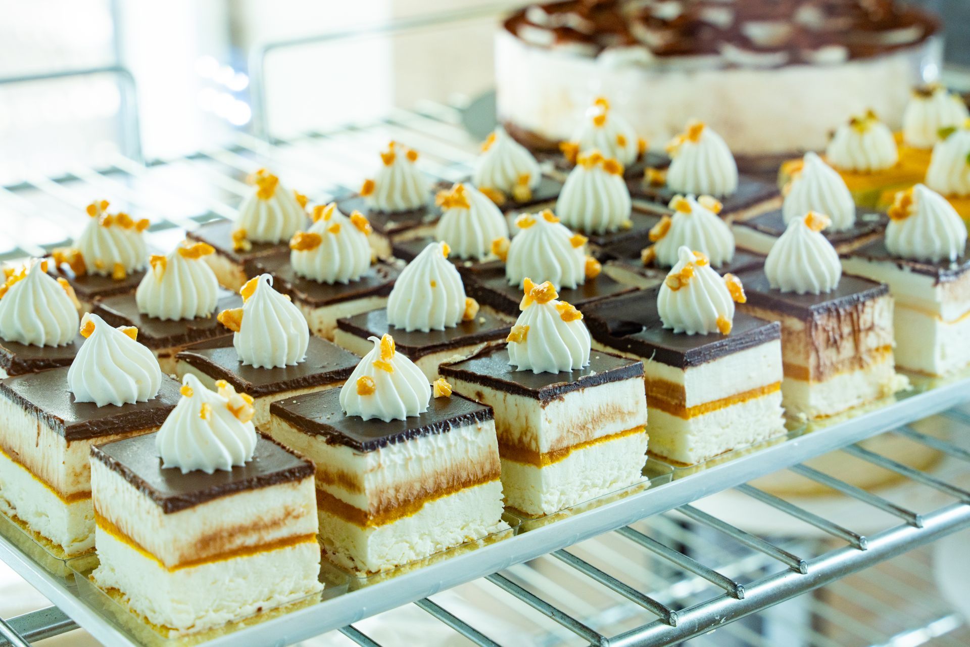 File di quadratini di dessert a strati, ricoperti di panna, su una griglia di raffreddamento, con una torta glassata sullo sfondo.