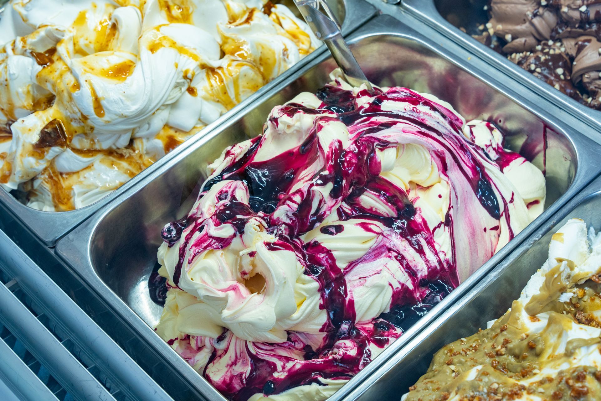 Assortimento di gusti di gelato in vassoi di metallo, tra cui bianco con caramello, rosa a spirale e giallo con copertura di briciole.
