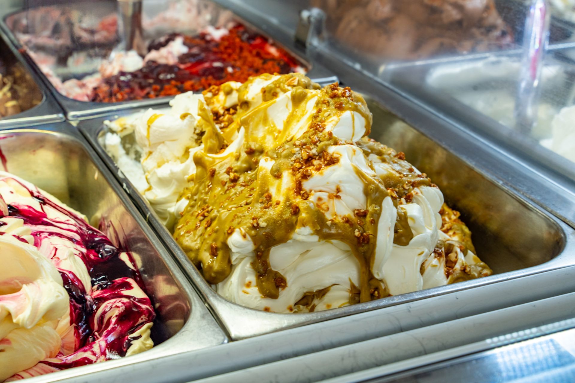 Assortimento di gusti di gelato in vassoi metallici, tra cui gelato bianco, giallo e ai frutti di bosco.