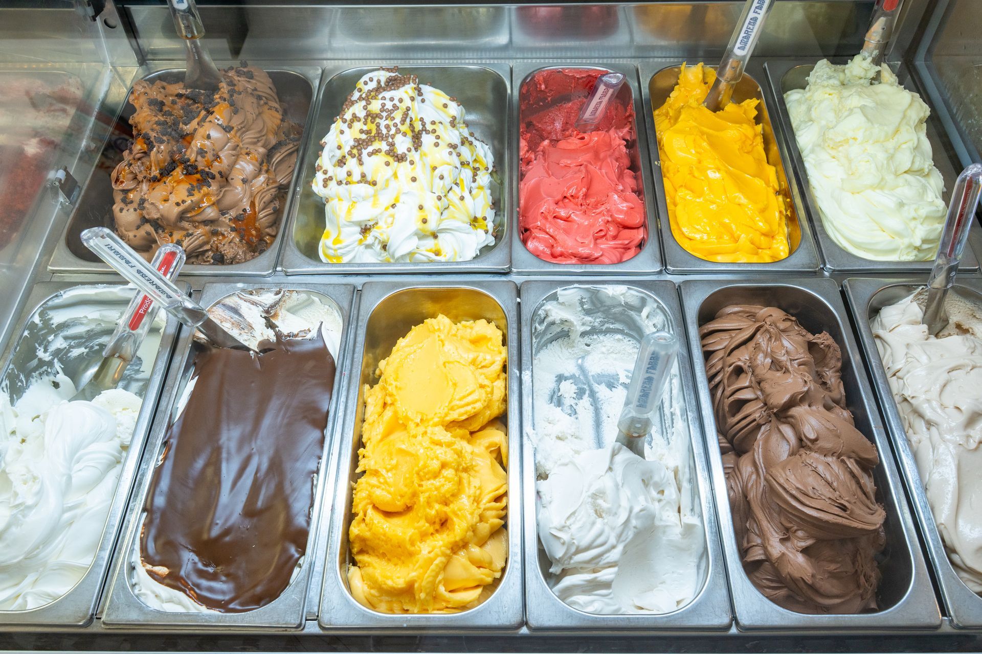 Assortimento di gusti di gelato in contenitori espositivi, tra cui palline di gelato bianco, al cioccolato, giallo, rosa e marrone.