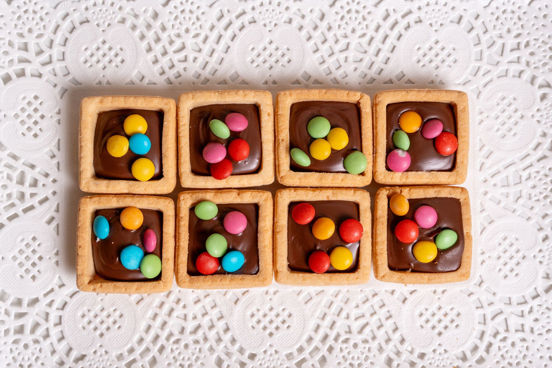 Otto piccole crostatine decorate con coloratissimi pezzi di caramelle, adagiate su una tovaglia di pizzo bianco.