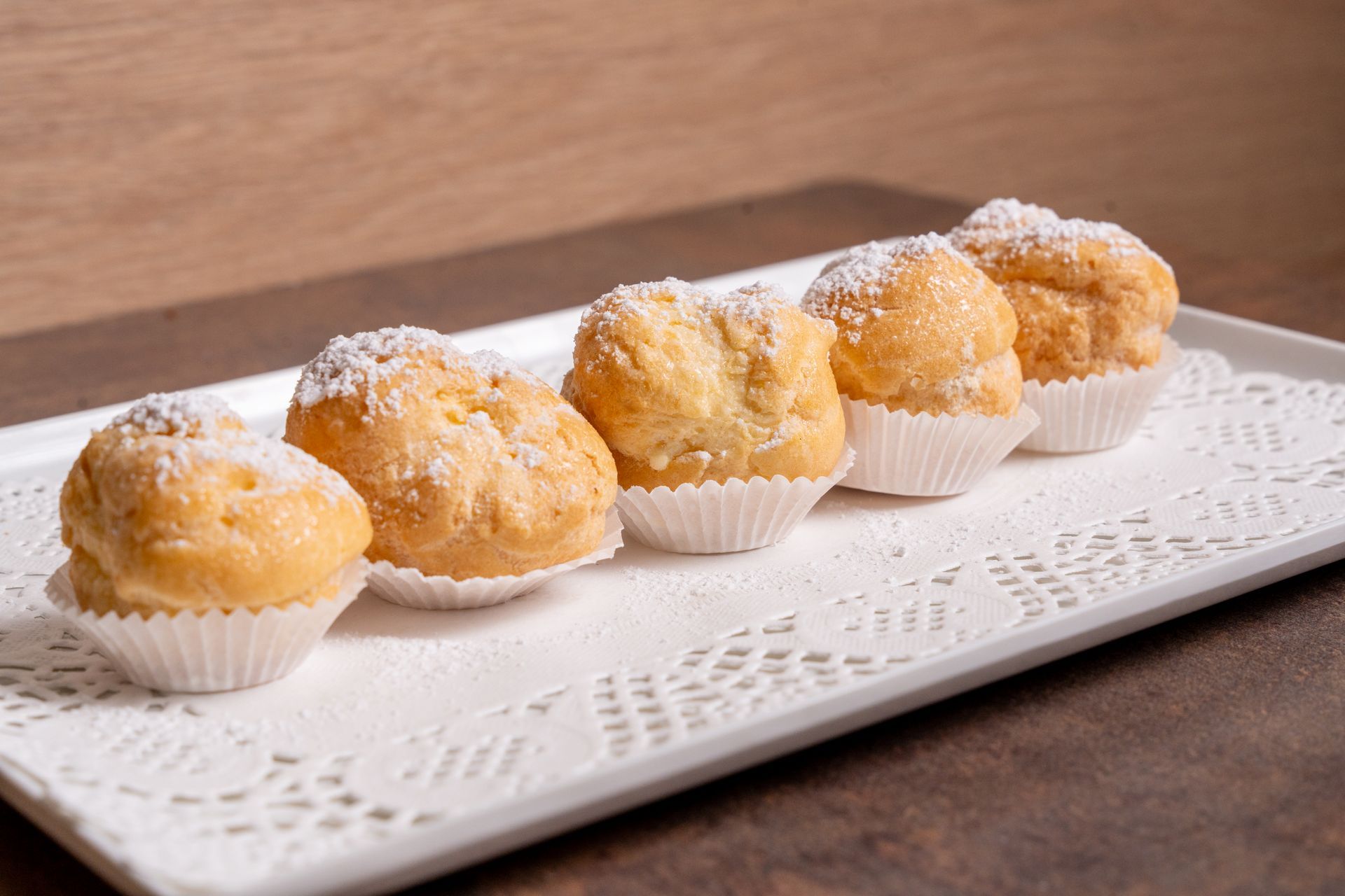 Cinque pasticcini ricoperti di zucchero a velo in pirottini di carta bianca su un vassoio bianco.