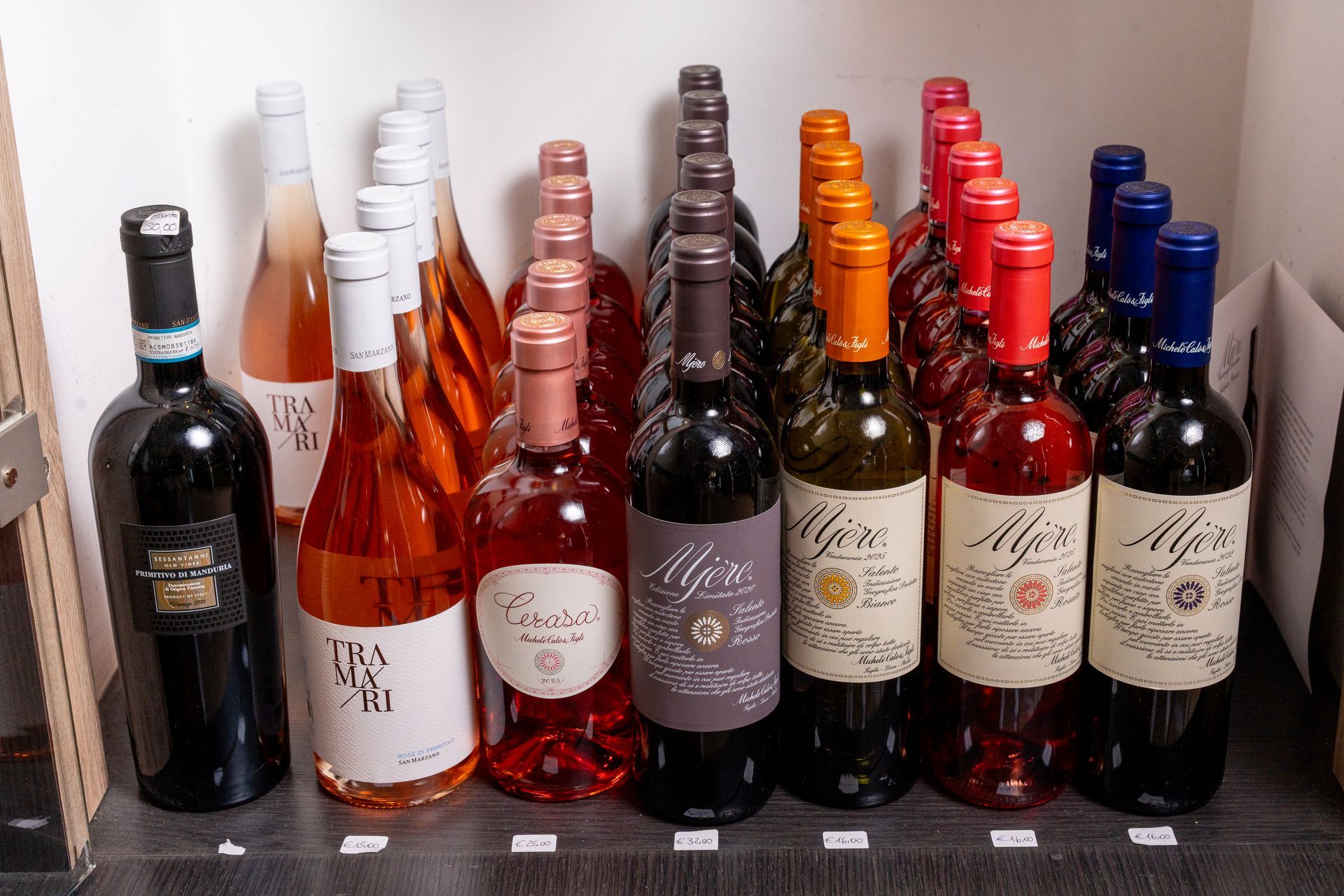 Diverse bottiglie di vino e spumante disposte su uno scaffale, etichettate per tipologia e colore.