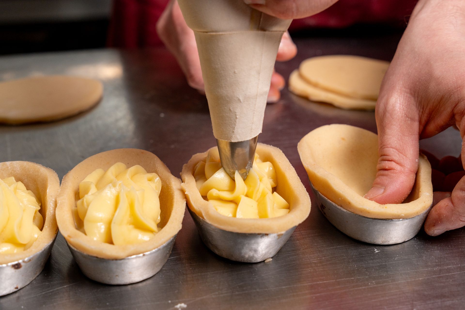 Mani che riempiono i gusci di pasta frolla in piccole crostatine su un vassoio di metallo.
