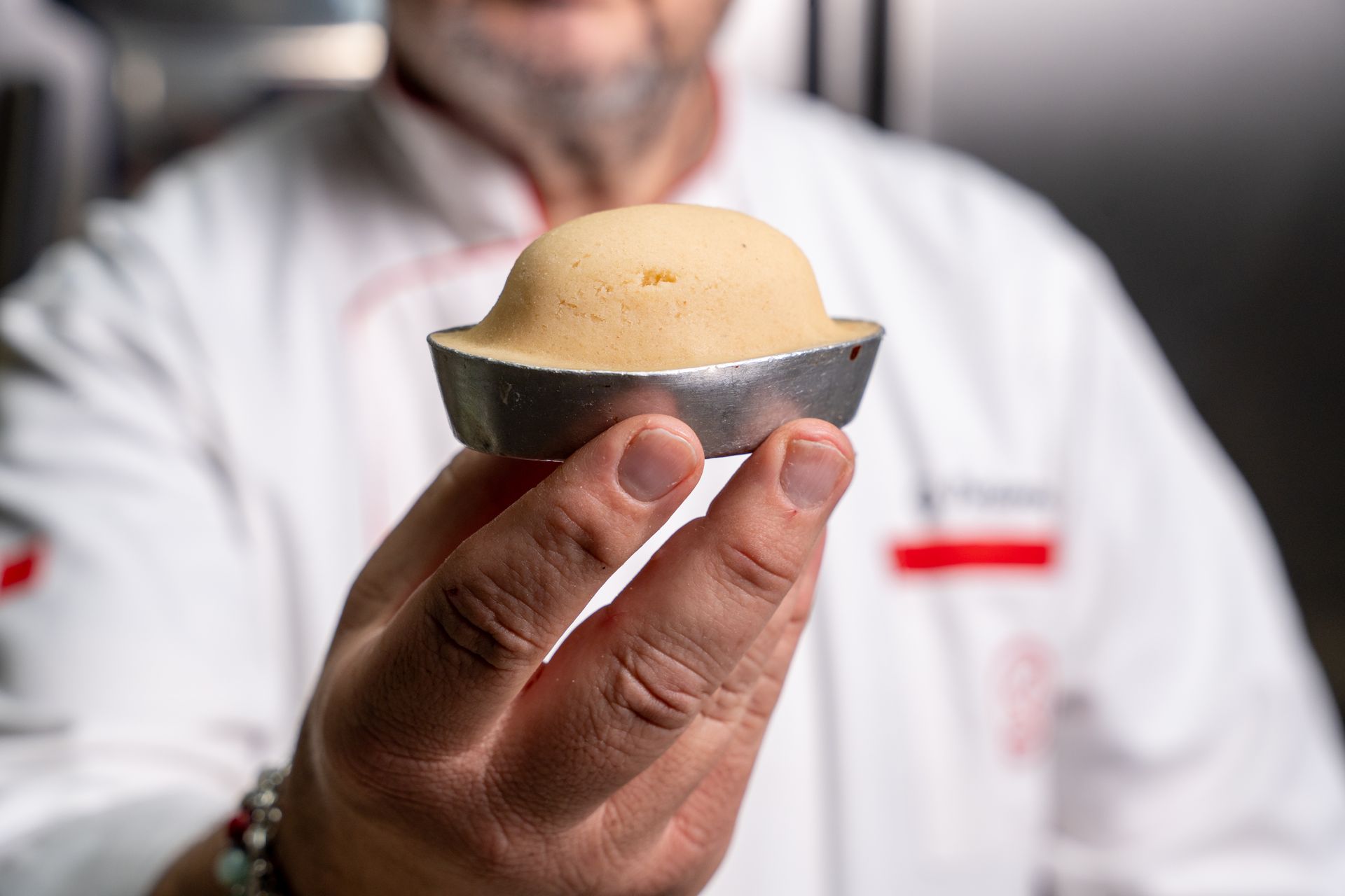 Una mano regge un piccolo dessert rotondo con base scura e parte superiore beige, mentre sullo sfondo si intravede una persona sfocata.