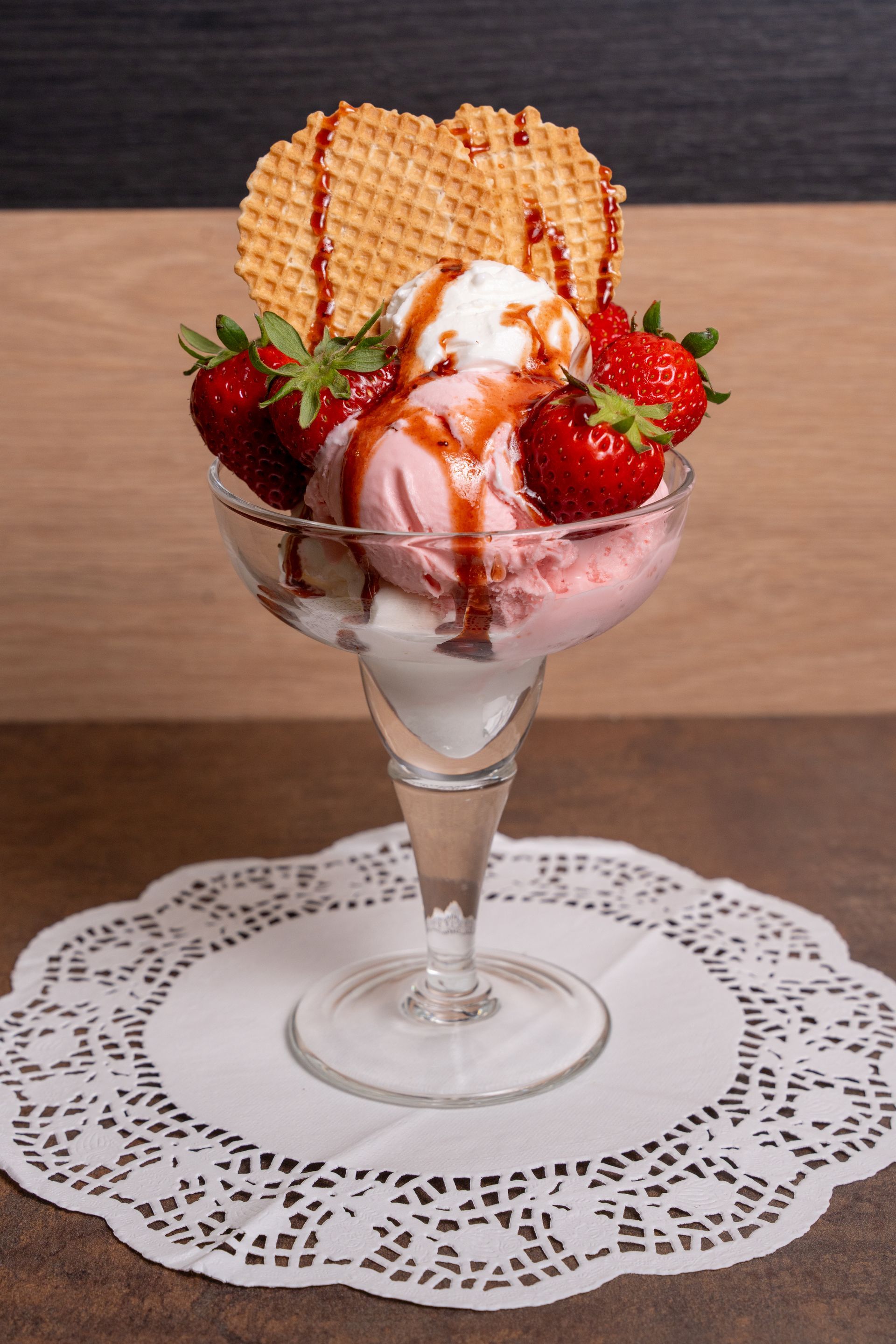 Coppa gelato alla fragola e vaniglia in un bicchiere con biscotti wafer, su un centrino di pizzo.
