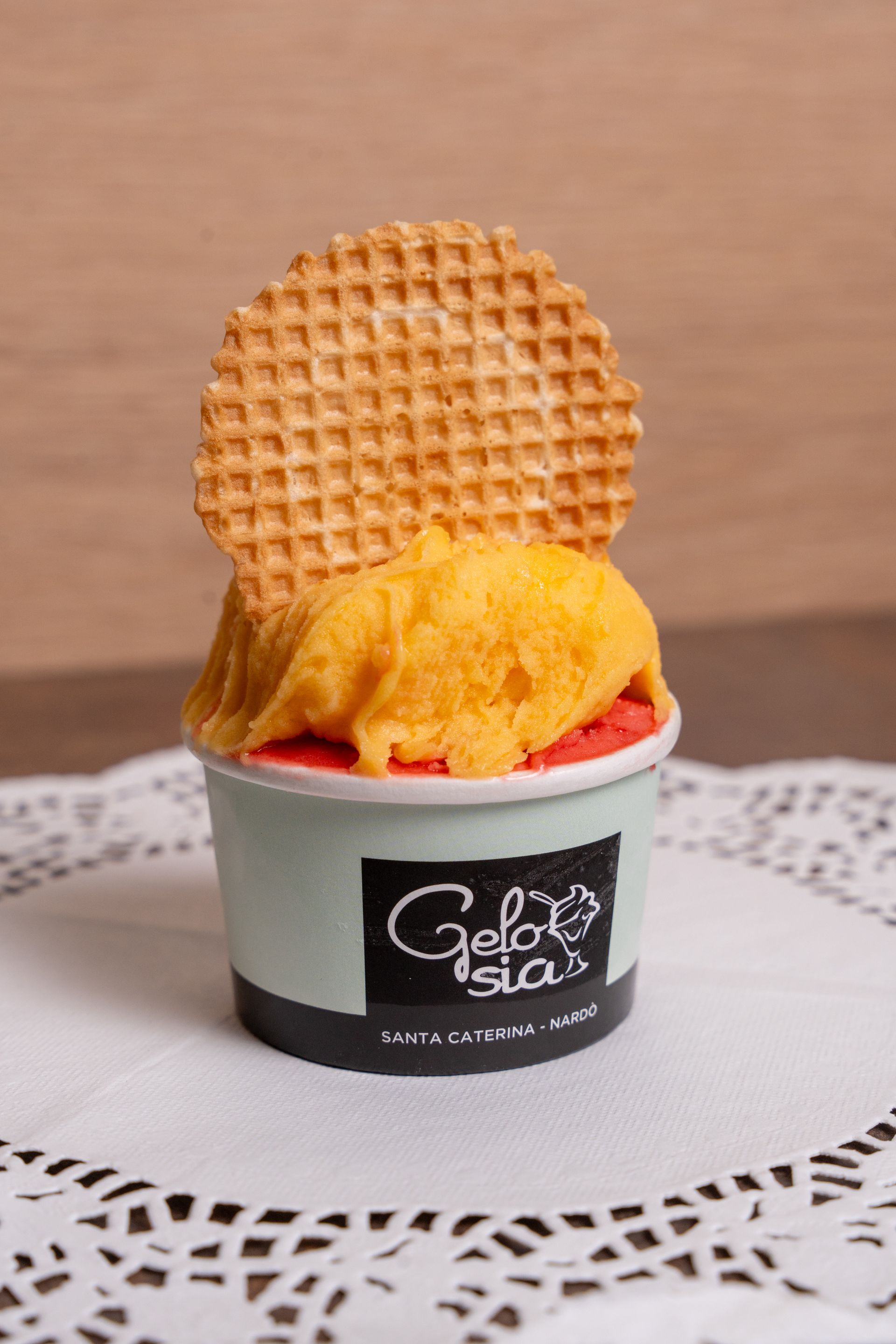 Gelato all'arancia in una coppetta nera con un biscotto waffle, su una superficie di pizzo.