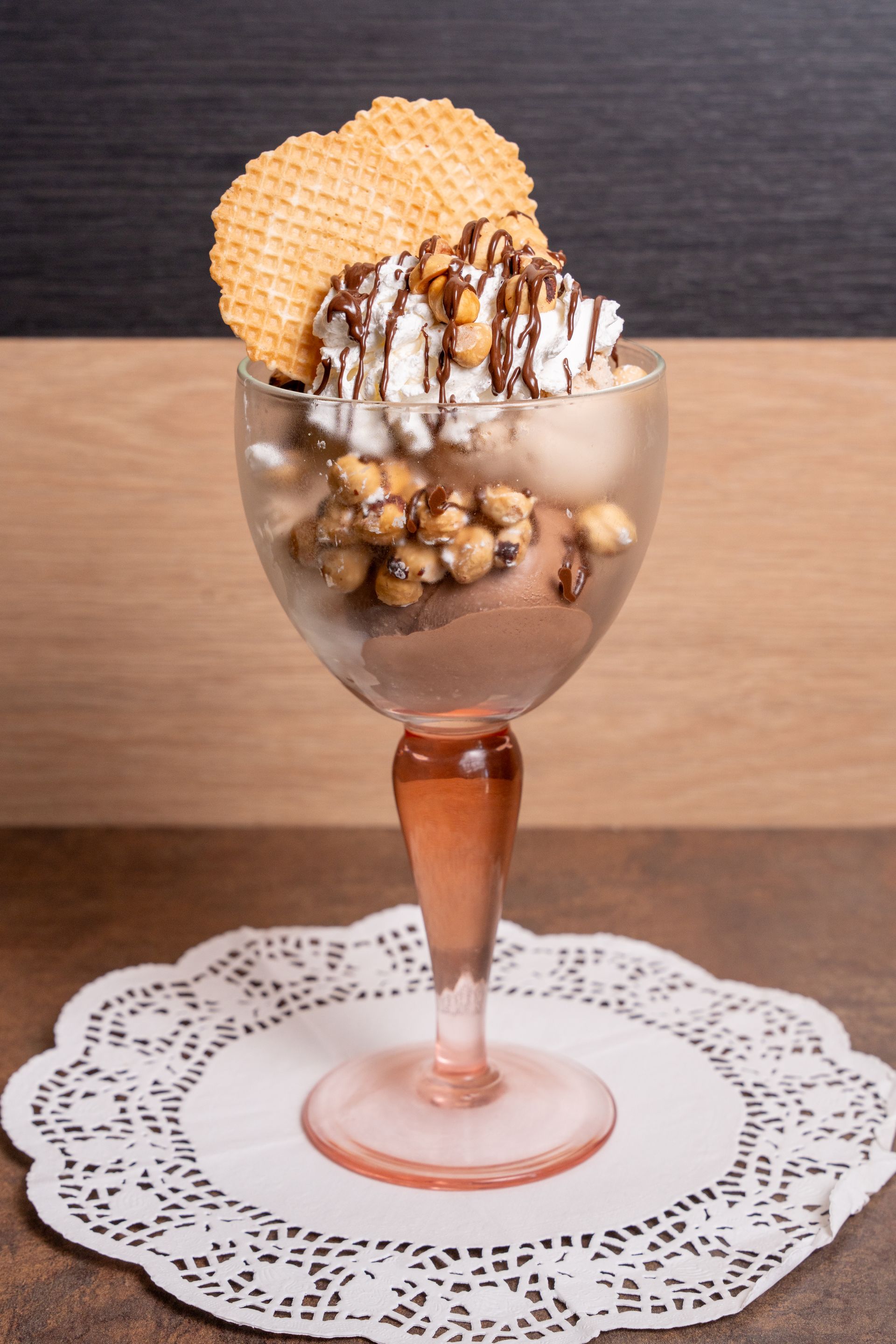 Coppa gelato in un bicchiere con stelo, guarnita con cialda e glassa al cioccolato su un centrino di pizzo.
