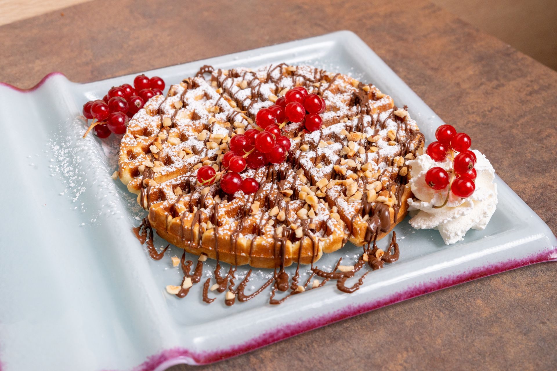 Waffle guarnito con panna montata, frutti rossi e zucchero a velo su un piatto bianco.