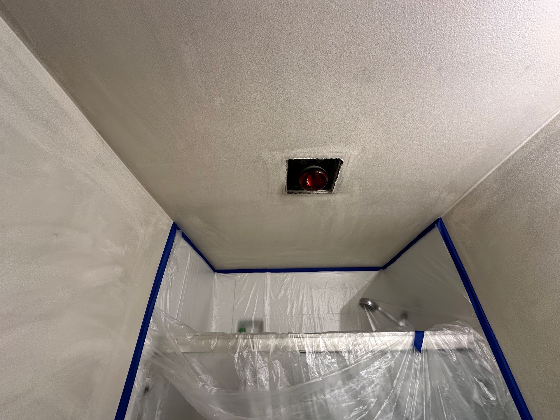 A bathroom ceiling with a rectangular cutout for a light fixture, protected by blue painter's tape and plastic sheeting.