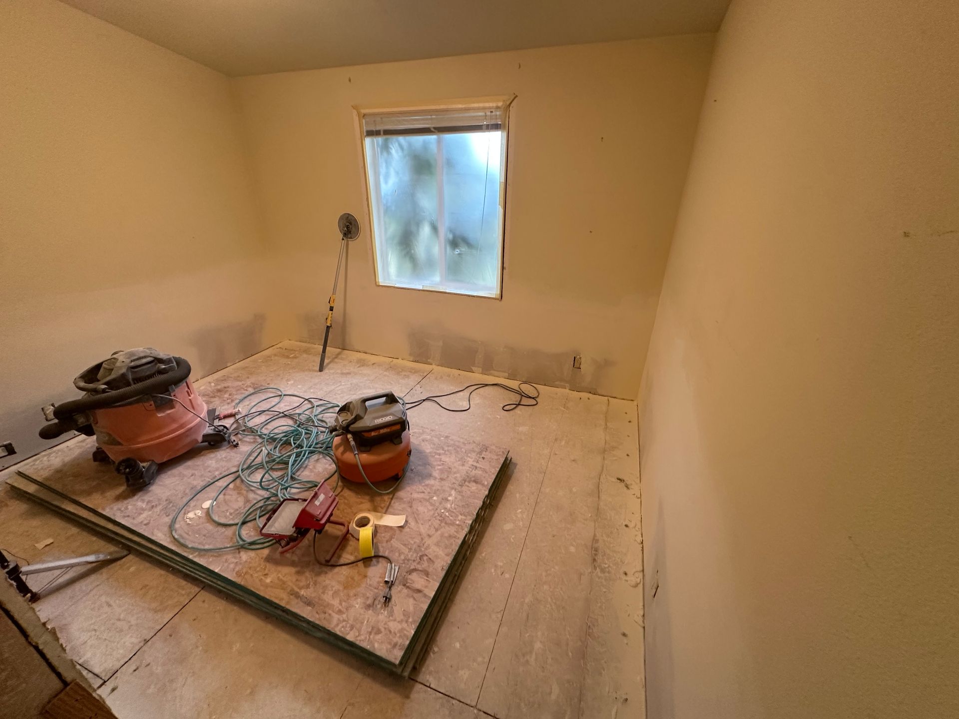 An unfinished bedroom with plywood subflooring, construction tools, and a shop vacuum, mid-renovation.