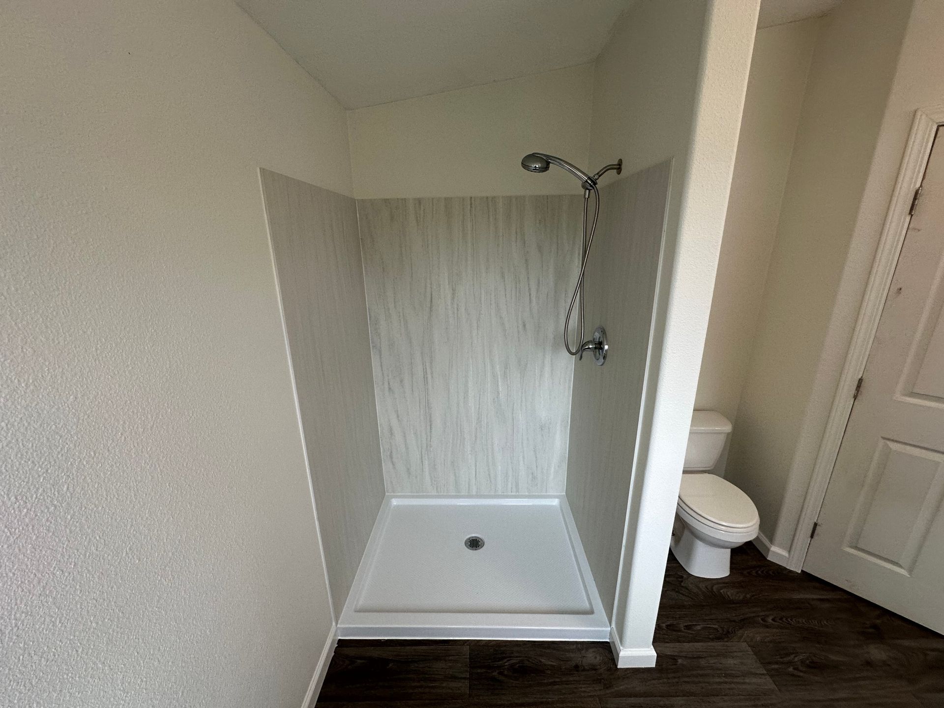 A white shower stall with a handheld shower head and textured wall tiling, next to a toilet in a bathroom with wood floors.