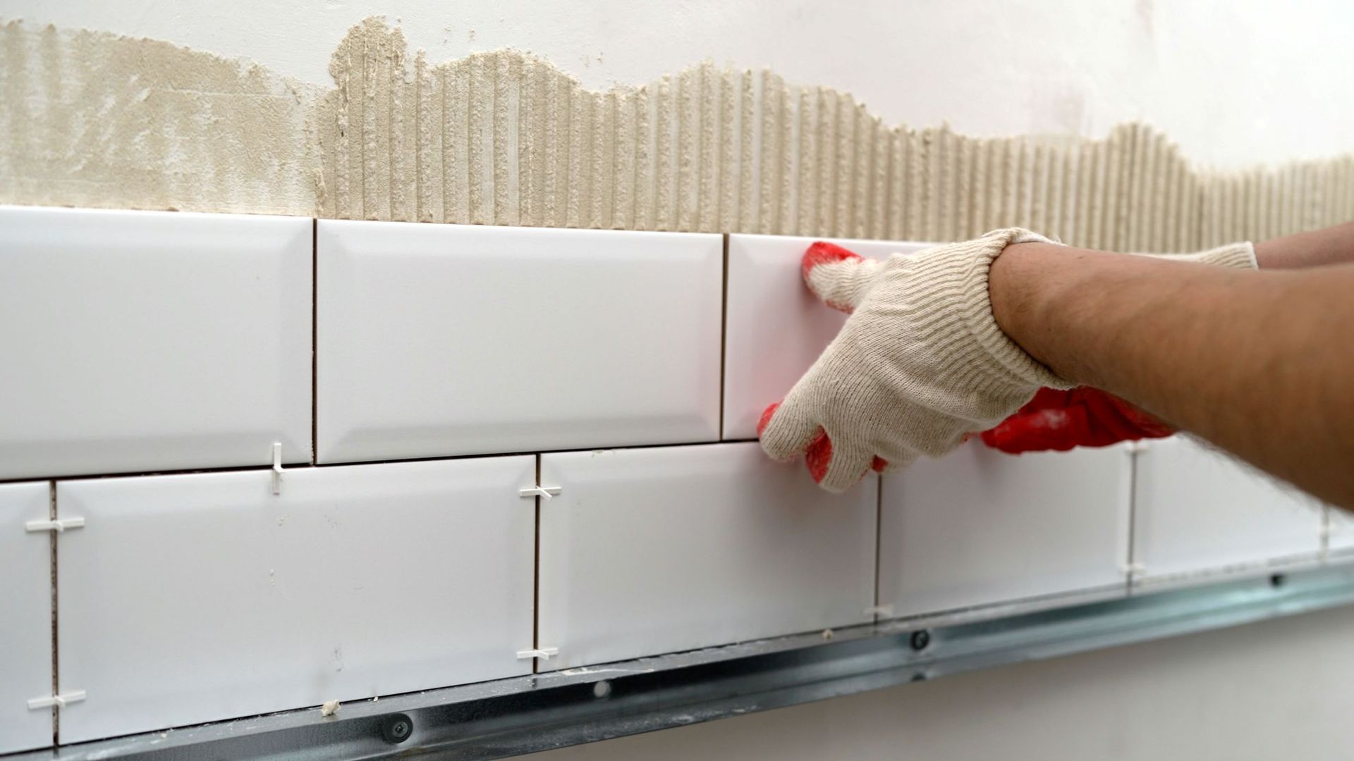 A gloved hand places a white subway tile onto a wall with combed adhesive, using spacers to align it with others.