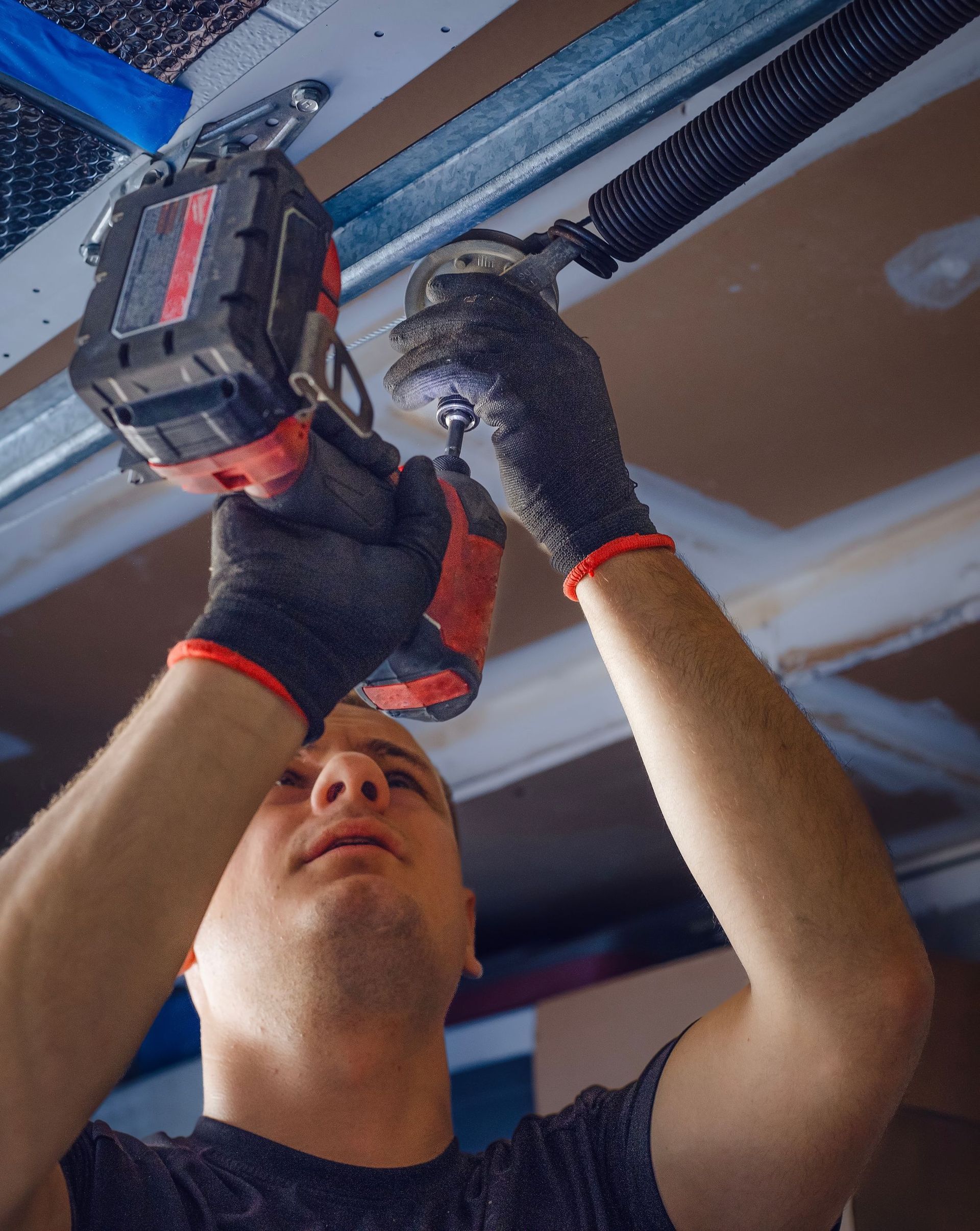 A person wearing work gloves uses a red power drill to work on a garage door torsion spring mounted on the ceiling.