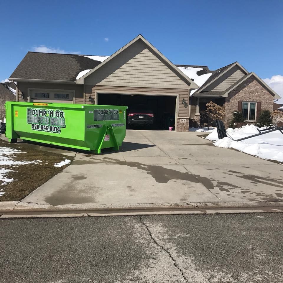 Green dumpster in front of a house, driveway with melting snow, blue sky.
