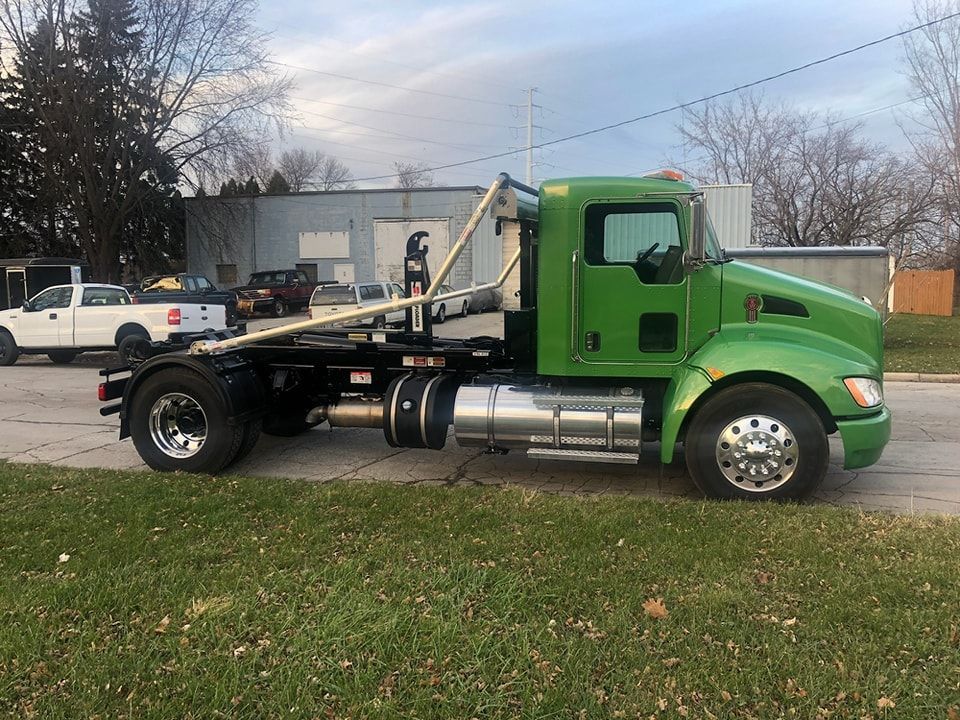 Green industrial truck on a street, with a white truck in the background. Cloudy sky.