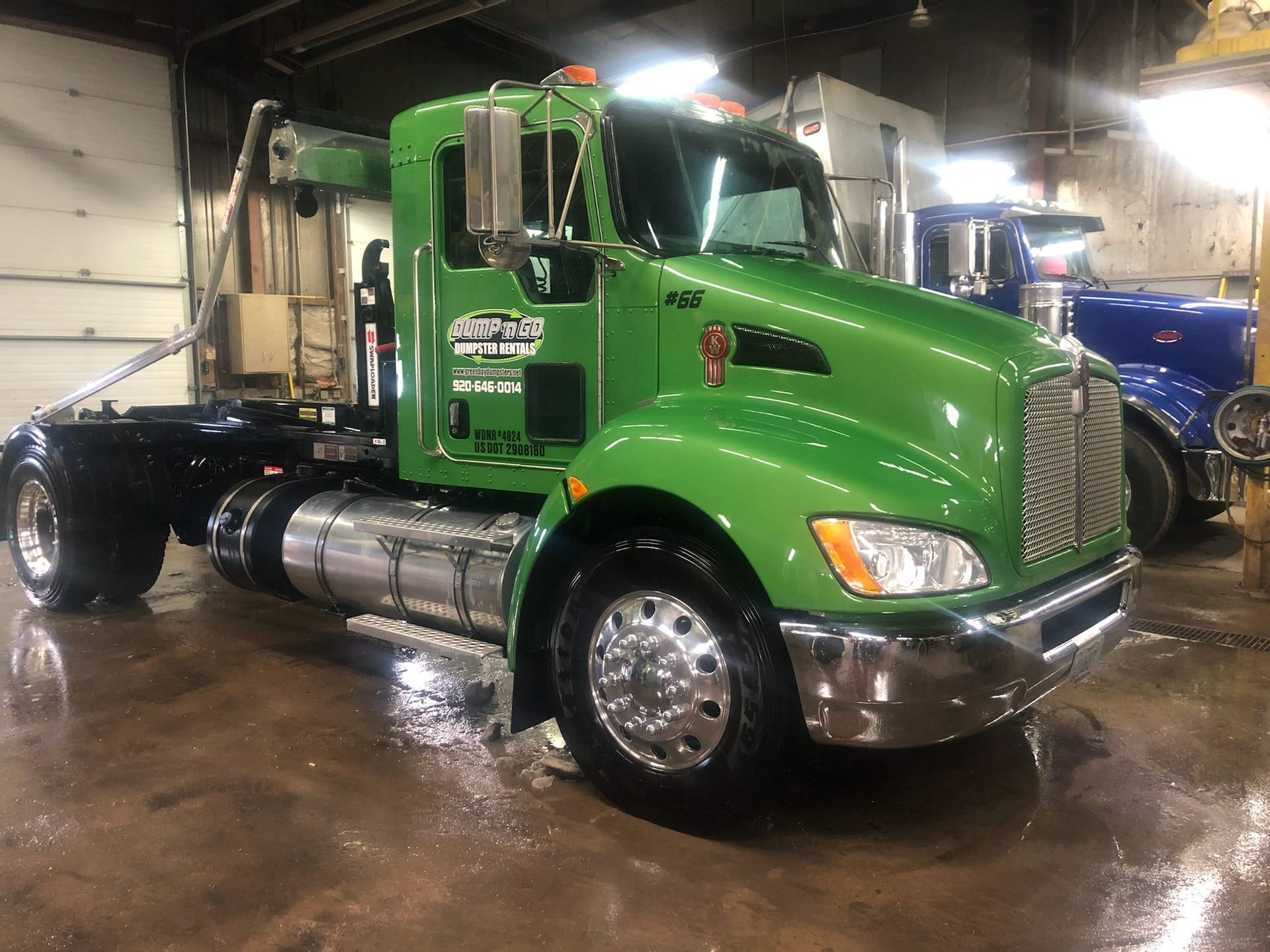 Green semi-truck, parked in garage. Shiny, chrome features, with black frame. Another truck in the background.