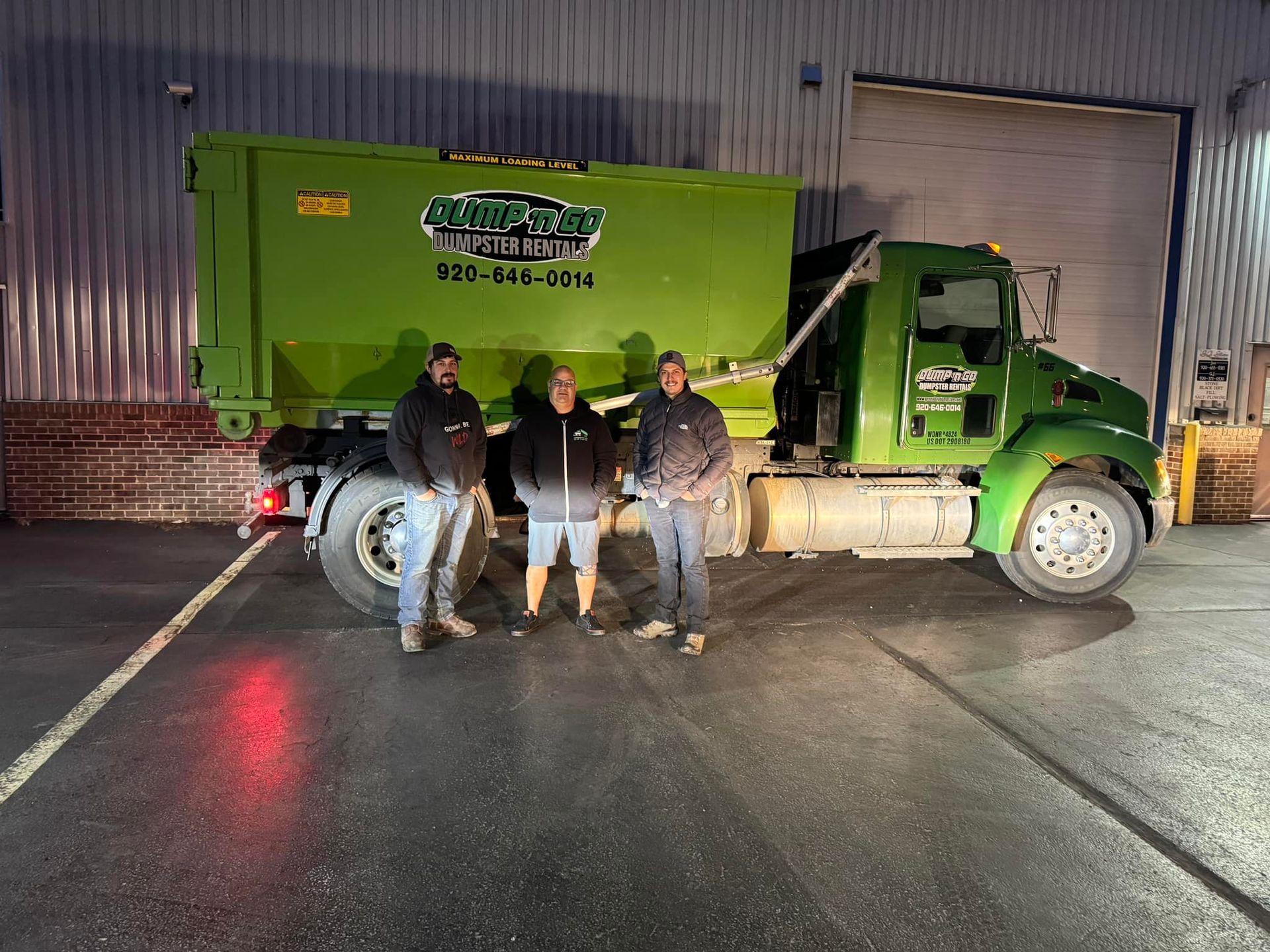 Three people standing by a green dumpster truck in front of a building.
