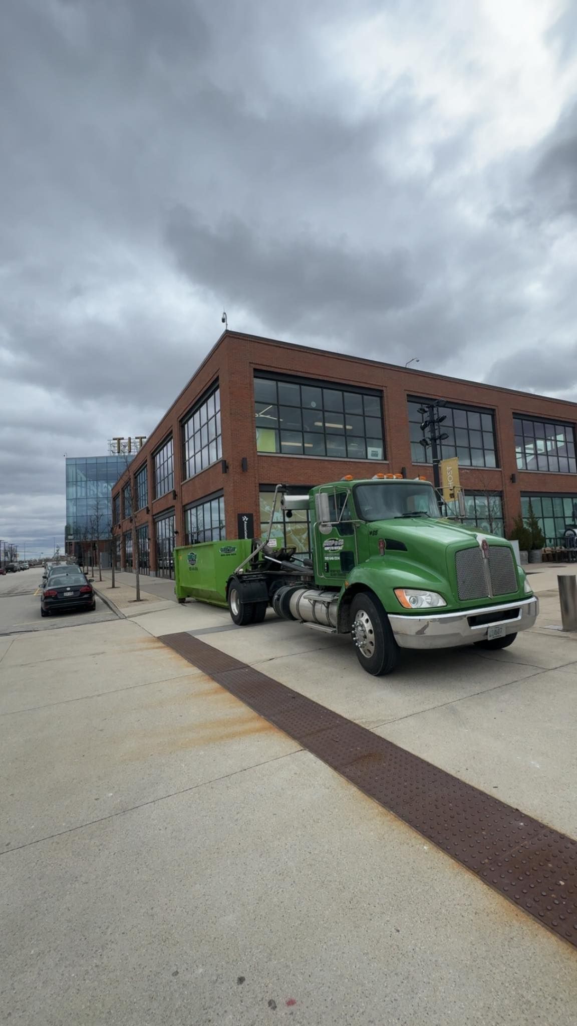 Green semi-truck towing a green container in front of a brick building on a cloudy day.
