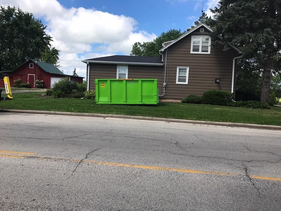 Green dumpster on grass in front of a brown house; a red barn is in the background.