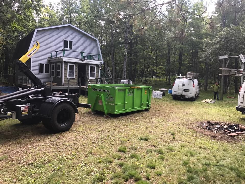 Green dumpster being loaded by a truck near a house, two vans, and trees.