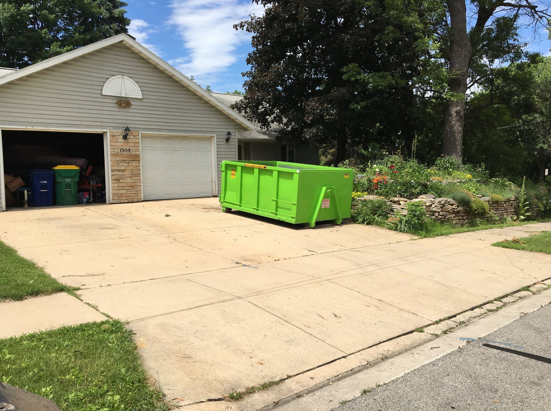 Green dumpster in driveway of a house with open garage. Blue and black trash bins are visible.