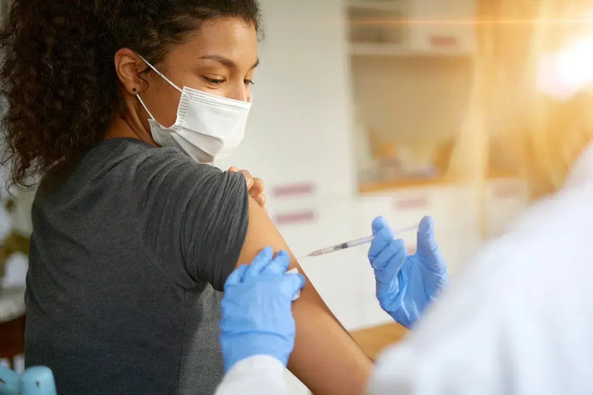 A woman wearing a mask is getting a vaccine in her arm. A woman wearing a mask is getting a vaccine in her arm.