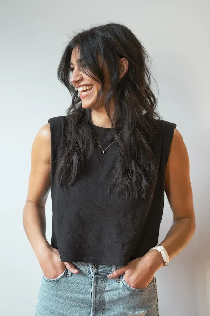 Woman with dark hair laughs, hands in pockets, wearing black top and jeans. Light background.