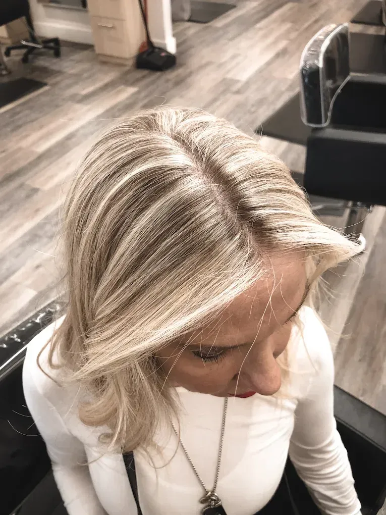 Blonde woman with parted hair, highlights visible, sitting in a salon chair.