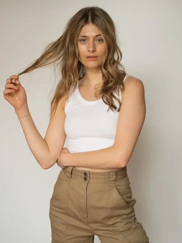 Woman in white tank top and khaki pants, holding hair, looking at the camera.