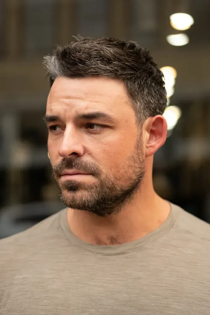 Man with short graying hair and beard, wearing a tan shirt, looking off to the side.