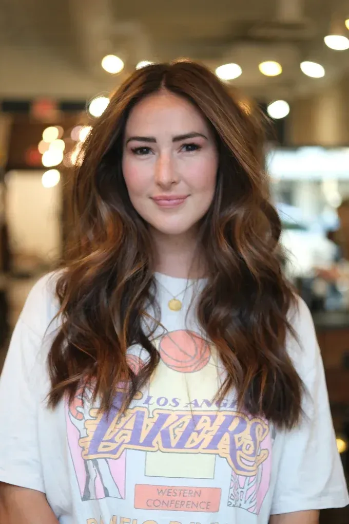 Woman with wavy brown hair, wearing a Lakers t-shirt, smiles in a salon.
