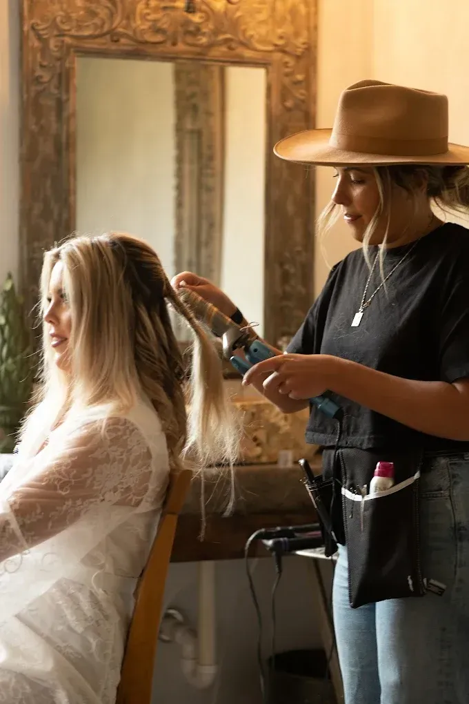 A hairstylist curls a woman's hair in a well-lit room, prepping for an event.