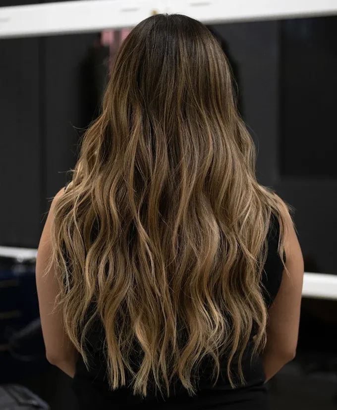 Woman with long, wavy, brown hair, highlighted with blonde, stands indoors.