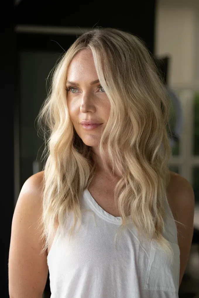 Blonde woman with wavy hair in a white tank top, looking off to the side.