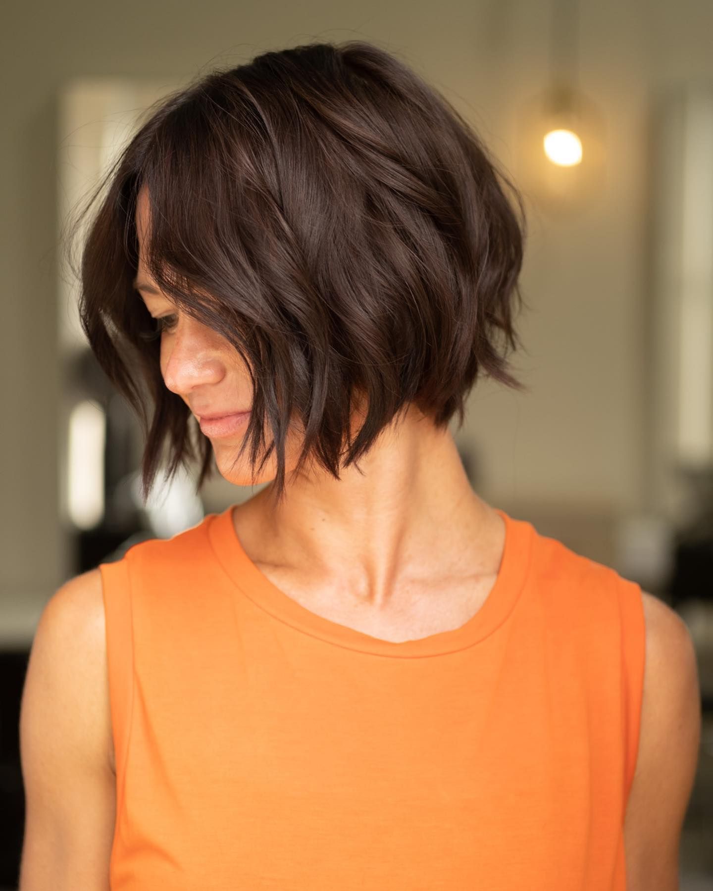 Woman with dark, wavy bob haircut, wearing orange top, looking down.