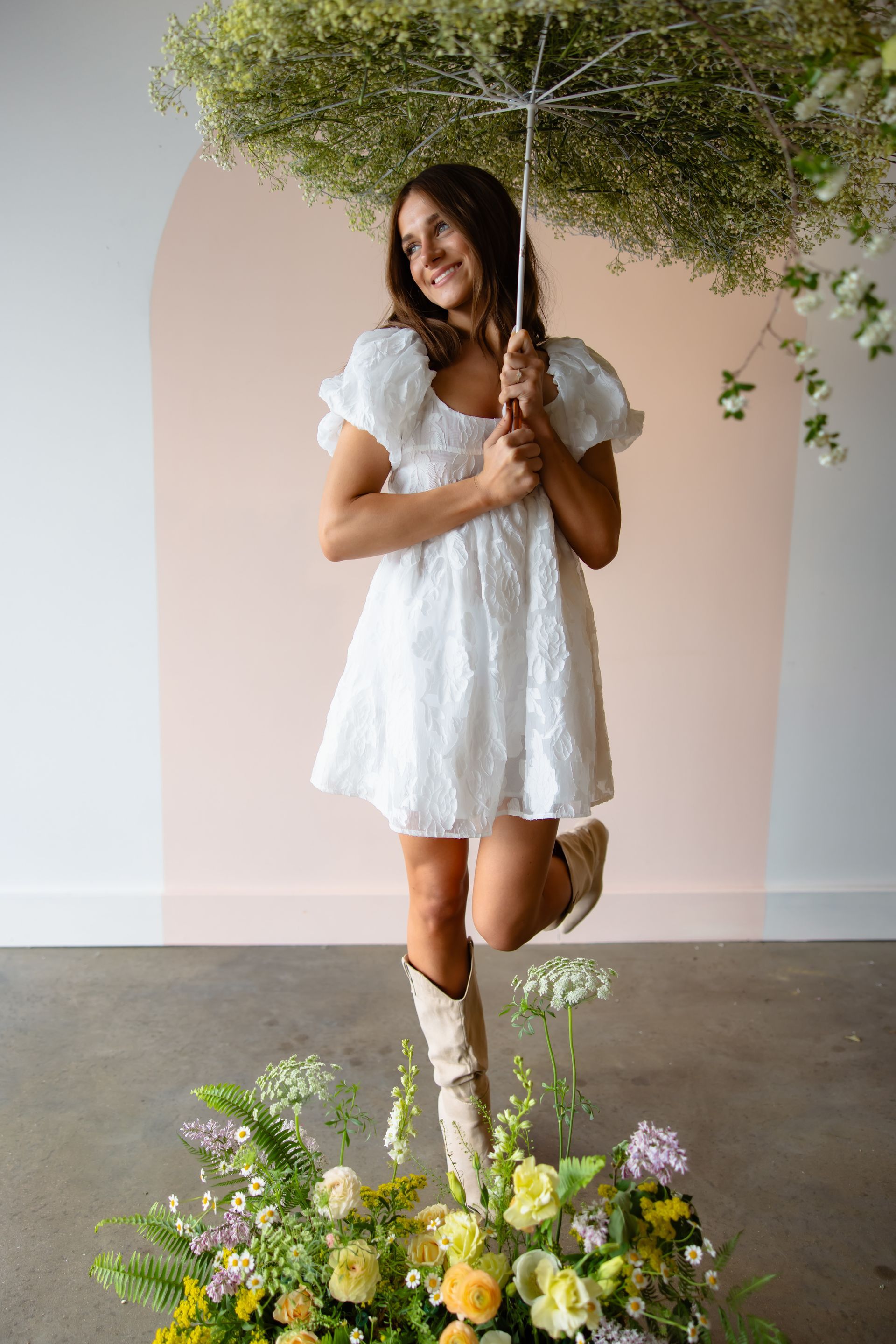 Woman in white dress holds floral umbrella, stands on floral arrangement, smiling. Pink and white wall background.