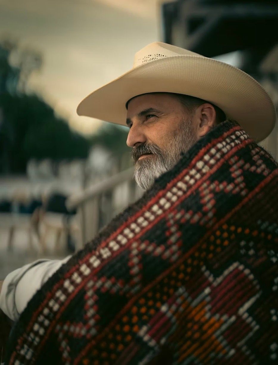Man in cowboy hat, wrapped in a patterned blanket, looking off to the side. Outdoor setting, muted colors.