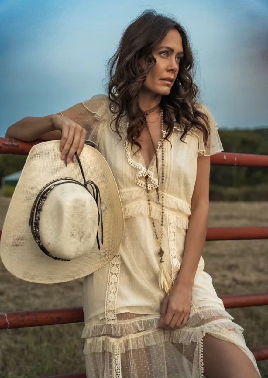 Woman in white dress leans on red fence, holding cowboy hat. Outdoors, thoughtful expression.