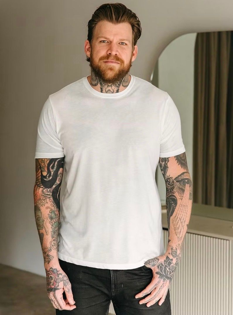 Man with tattoos wearing a white t-shirt and dark jeans, standing indoors with a neutral background.