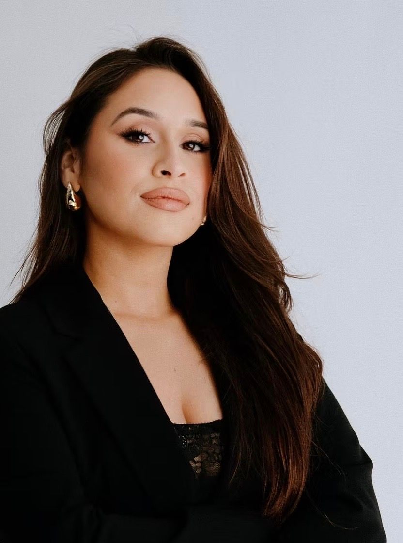 Woman with long brown hair, wearing a black blazer and earrings, looking at the camera.