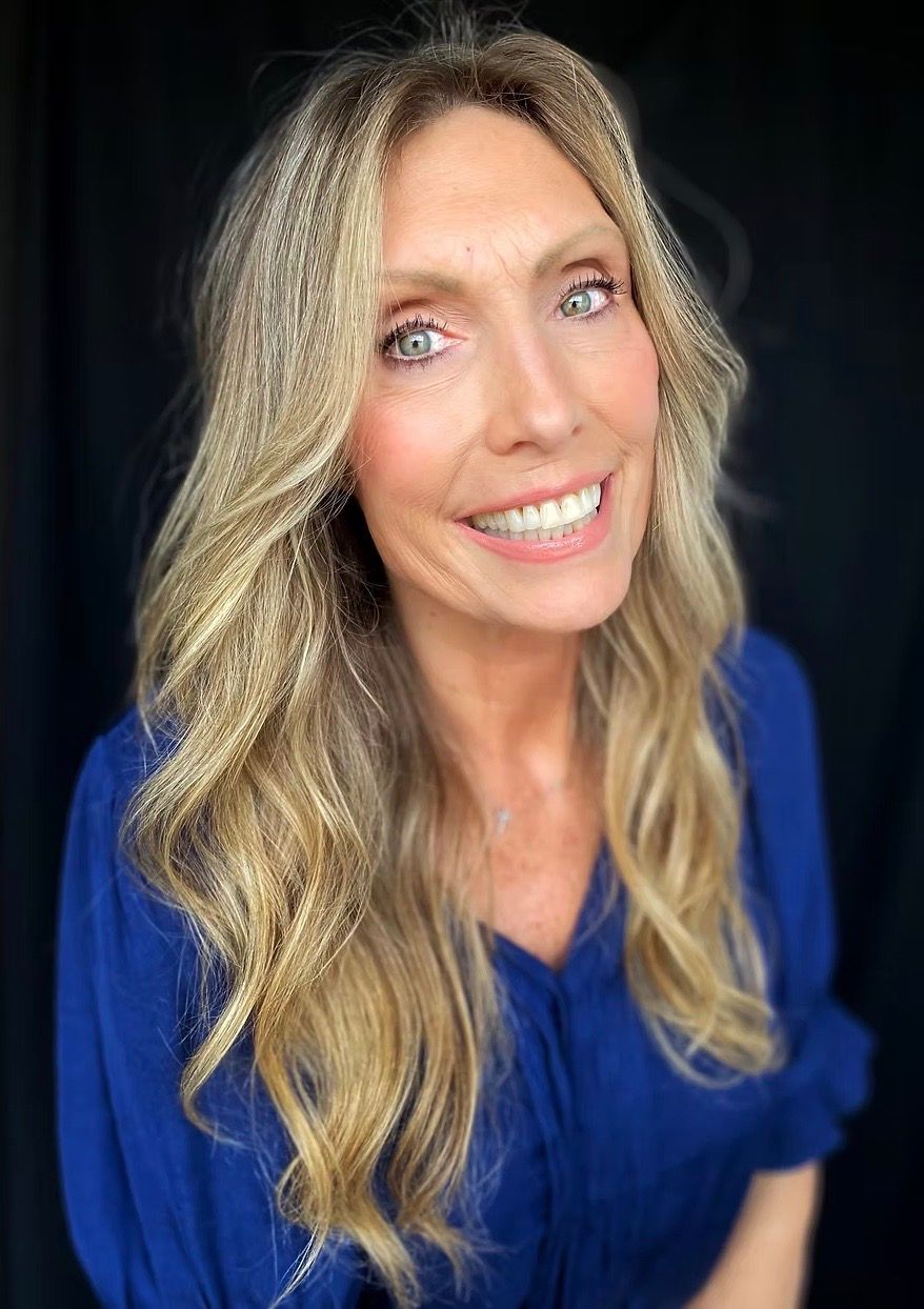 Blonde woman with blue eyes smiles. Wears blue top, hair has sparkle accents. Black background.