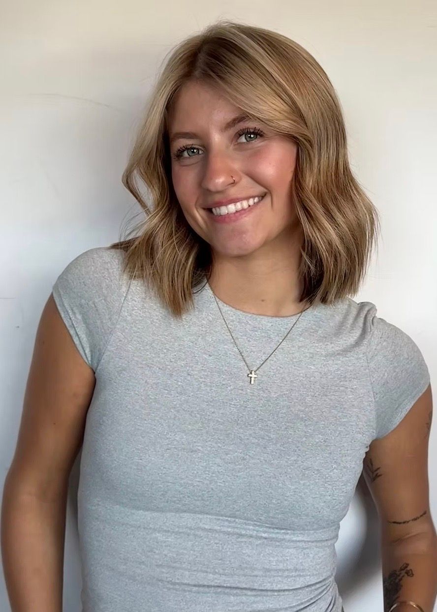 Young woman with blonde wavy hair smiles, wearing a gray top, small silver necklace, indoors.